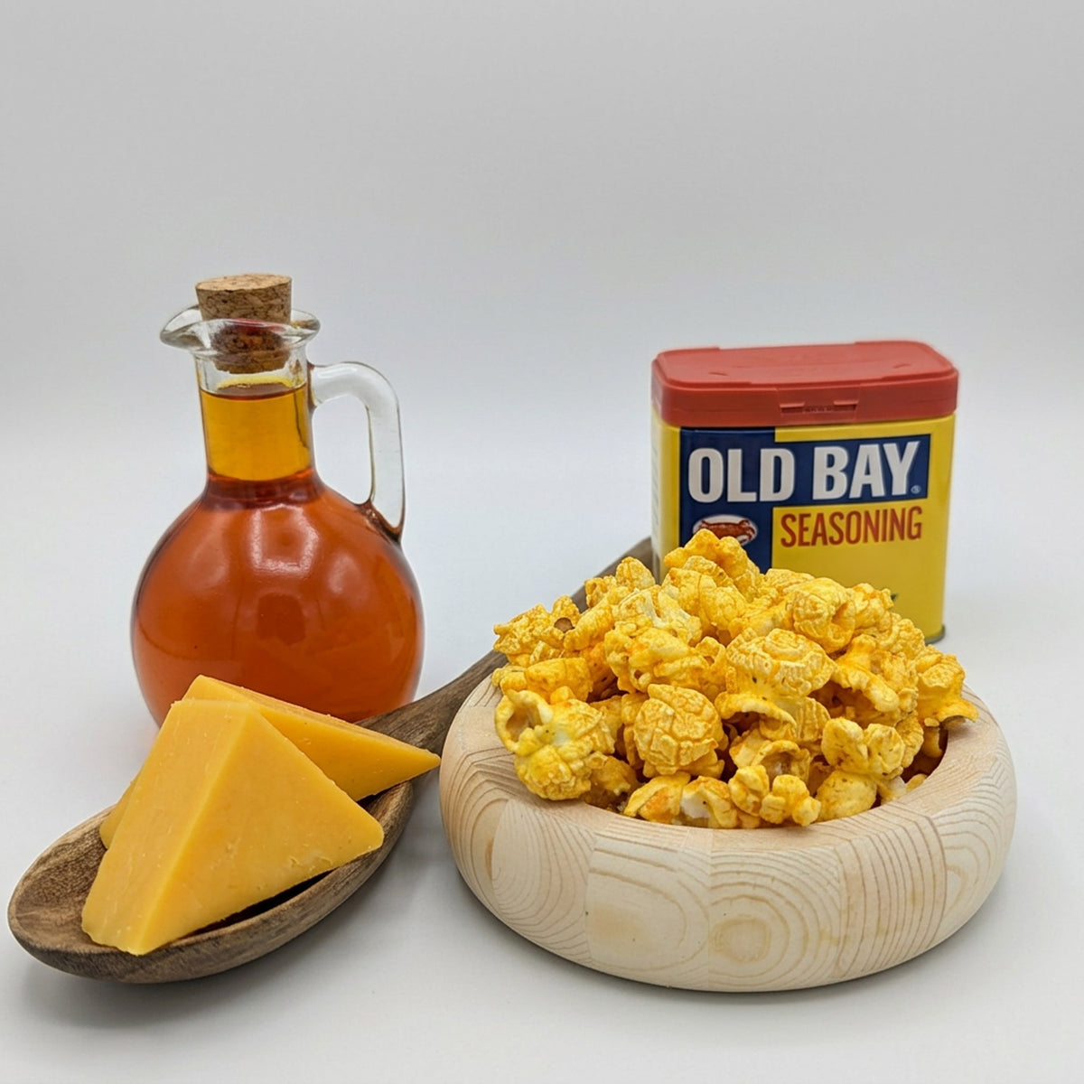 Bowl of popcorn, bottle of oil, and container of Old Bay seasoning on a white background