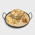 Glass bowl of popcorn on a wooden tray with handles, surrounded by cheese and caramels.