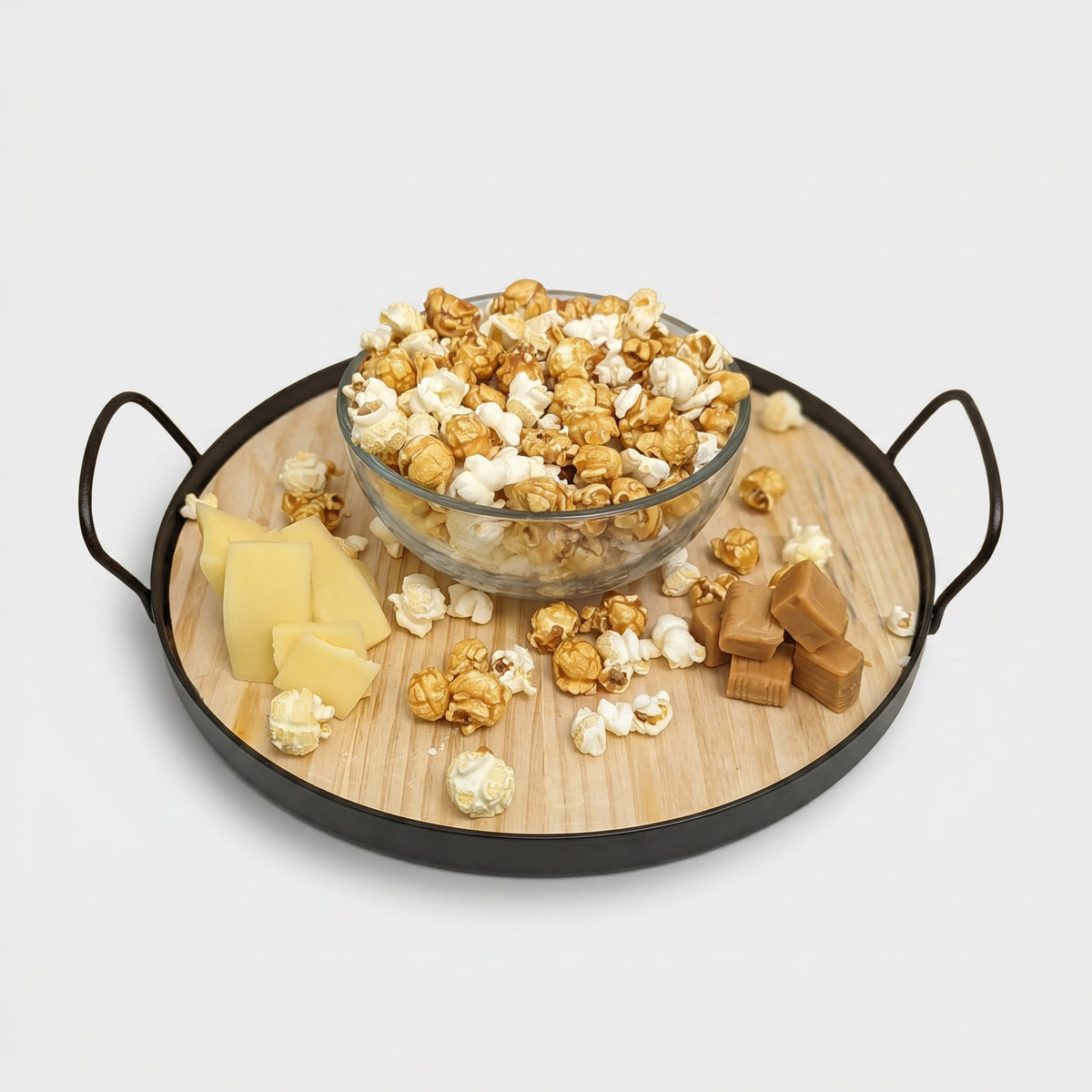 Glass bowl of popcorn on a wooden tray with handles, surrounded by cheese and caramels.