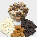 Glass bowl of chocolate-covered popcorn with white and dark chocolate pieces, caramels, and salt on a white background.