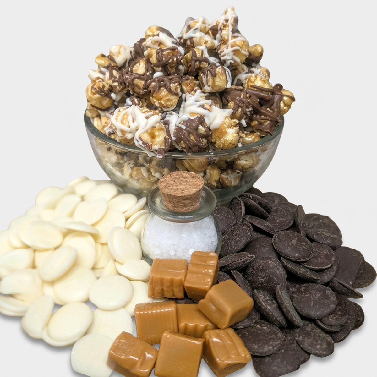 Glass bowl of chocolate-covered popcorn with white and dark chocolate pieces, caramels, and salt on a white background.