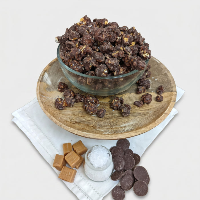 Glass bowl of chocolate-covered treats on a wooden plate with salt, caramels, and chocolate pieces.