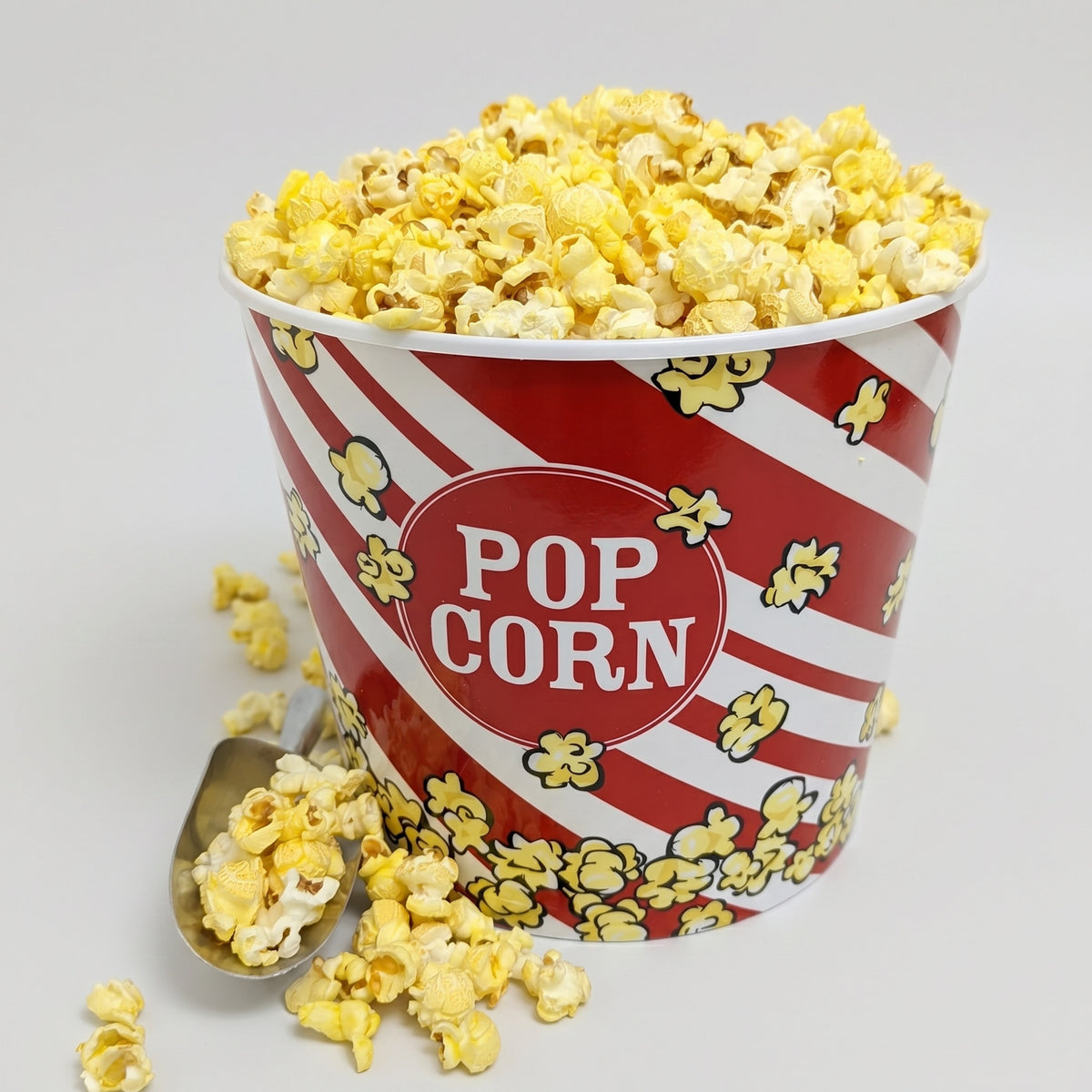 Red and white striped popcorn bucket with yellow popcorn and a spoon on a light gray background