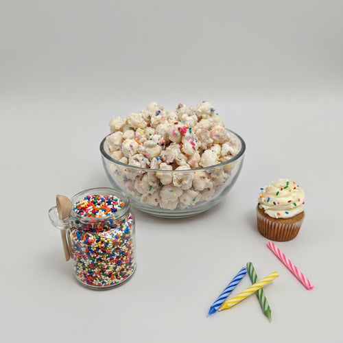 Glass bowl of popcorn with colorful sprinkles, jar of sprinkles, cupcake with sprinkles, and birthday candles on a light gray background.