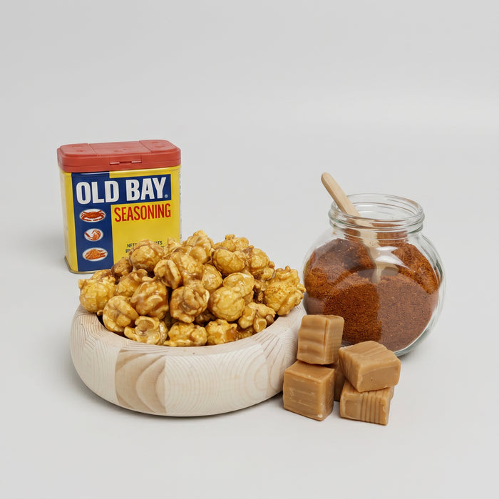 Bowl of popcorn, jar of brown sugar, and container of Old Bay seasoning on a light gray background