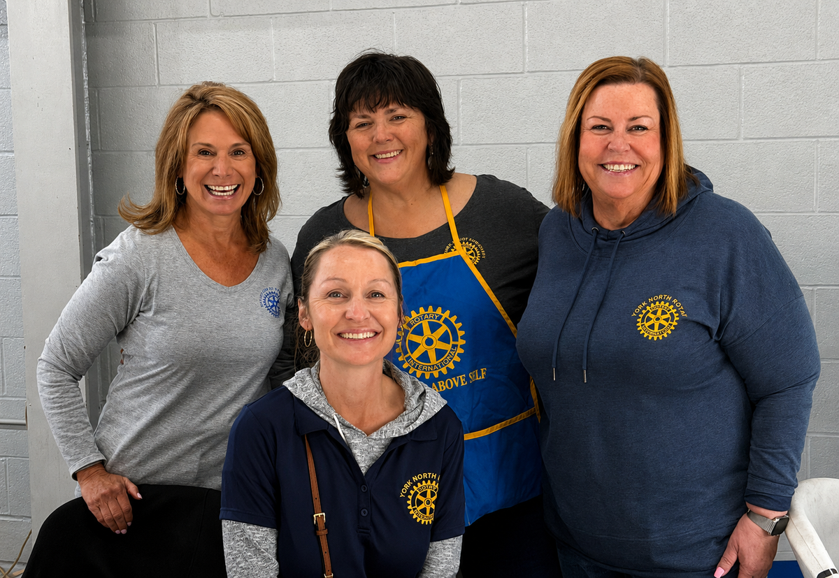 Four smiling women standing together in front of a light gray cinderblock wall, wearing matching navy blue Rotary International hoodies and a blue apron with yellow trim and logo.