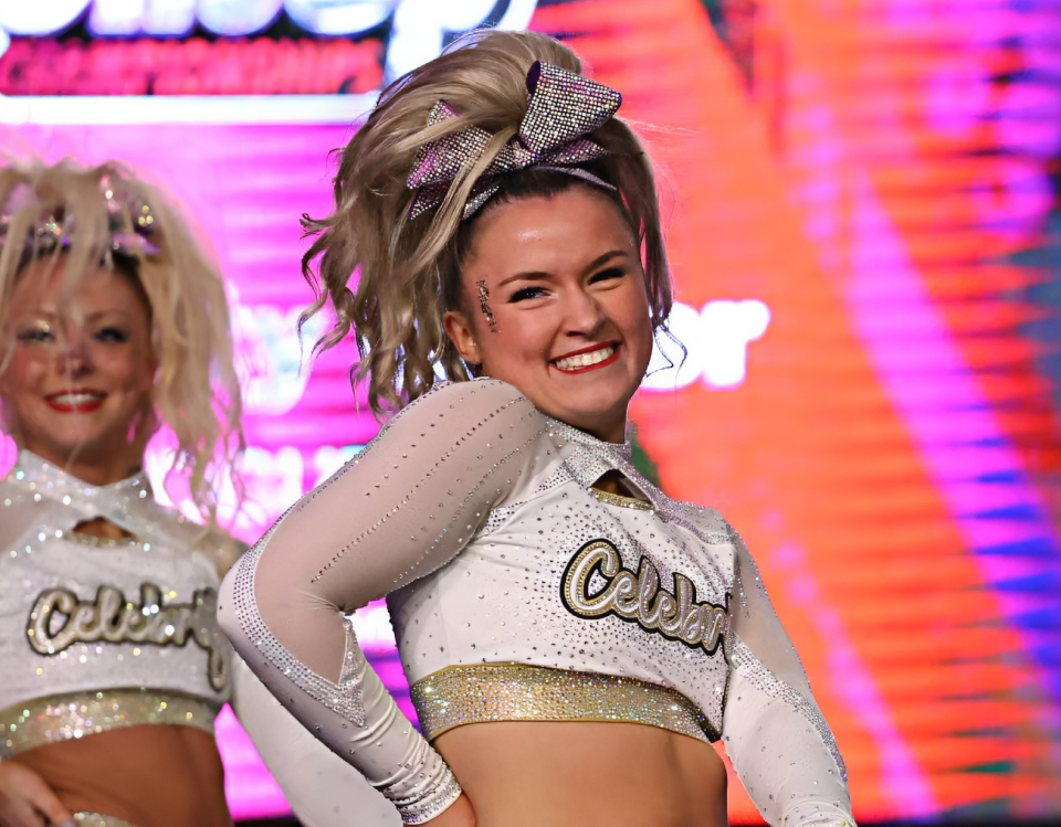 A cheerleader performer in a white and silver rhinestone crop top with 'Cotton' written across the front, posing on stage in front of a bright pink and orange lit background. Another cheerleader is partially visible in the background.