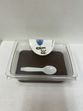 A square clear plastic container holding a dark brown dessert or pudding, with a small white plastic spoon resting inside and a white paper label on top.
