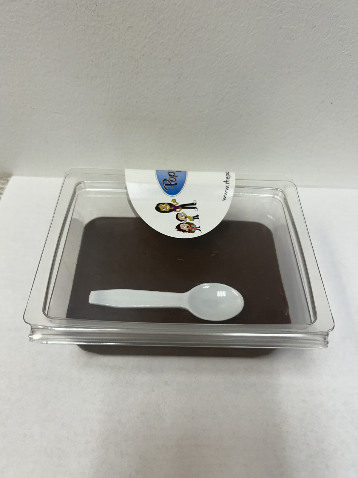 A square clear plastic container holding a dark brown dessert or pudding, with a small white plastic spoon resting inside and a white paper label on top.