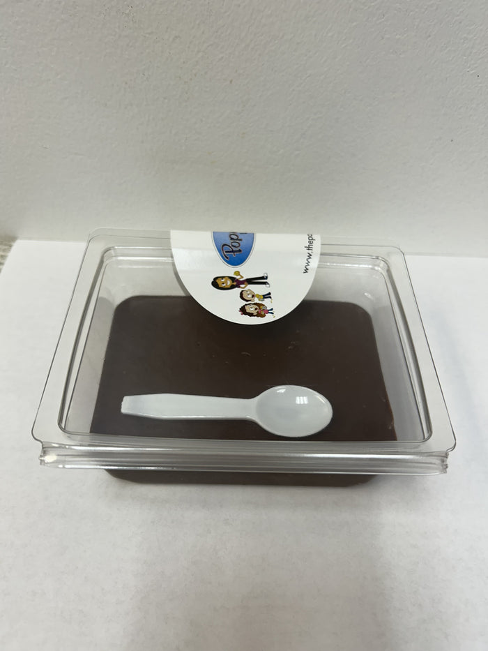 A square clear plastic container holding a dark brown dessert or pudding, with a small white plastic spoon resting inside and a white paper label on top.
