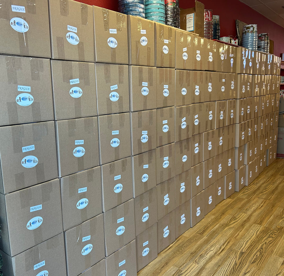 A long row of stacked brown cardboard boxes with white circular labels, photographed in a retail or storage room with wood flooring and shelves in the background.