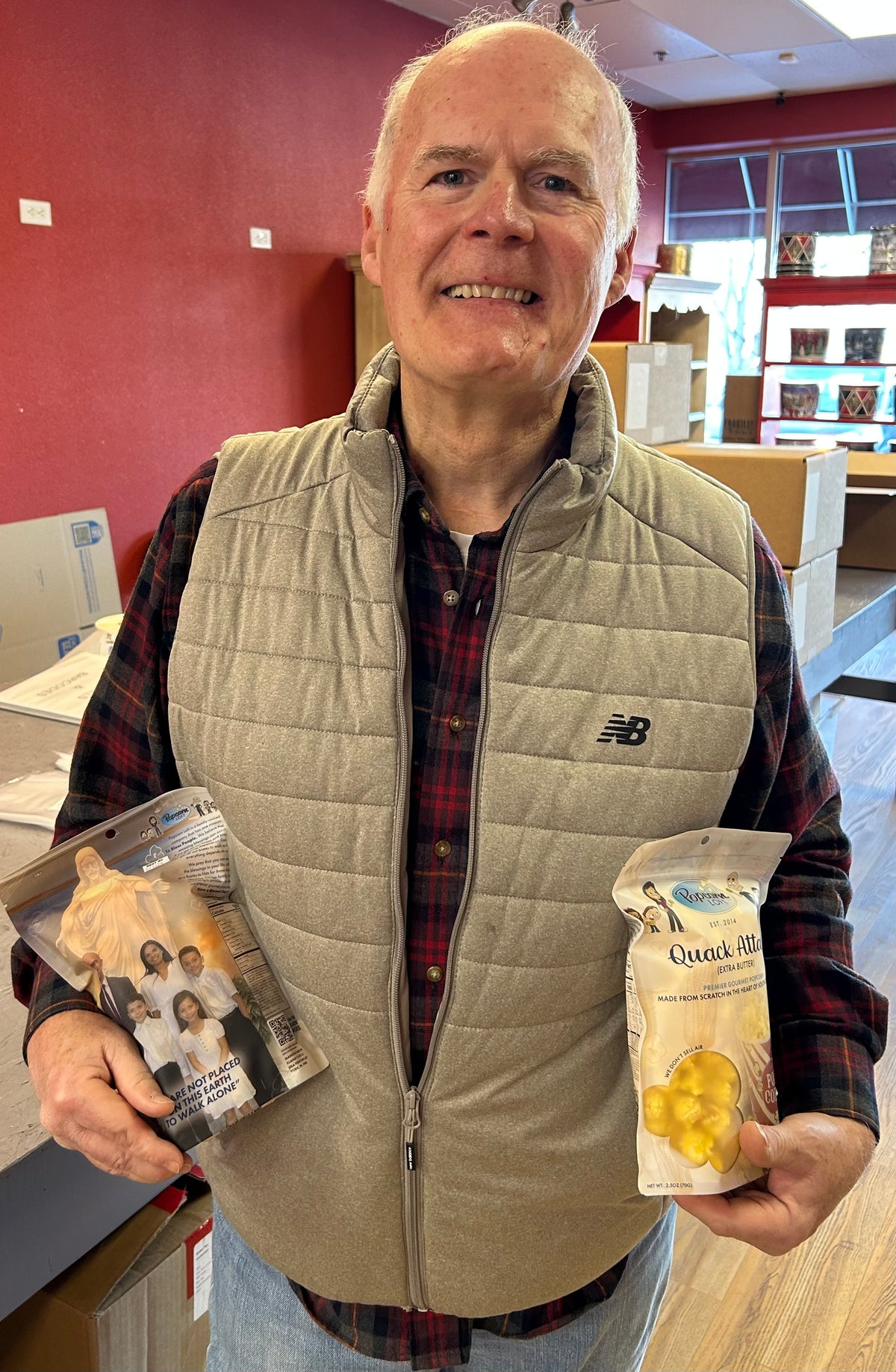 An older man in a gray vest holds two snack packages in a room with red walls and stacked boxes behind him
