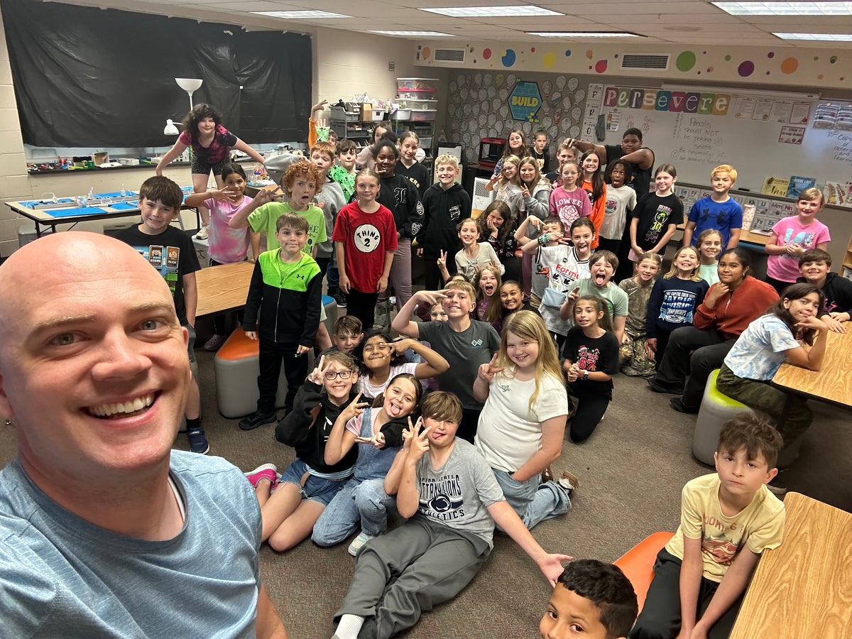 A large group of children and one adult man smiling and posing together in a classroom or school setting, photographed from an overhead angle with the photo rotated sideways.