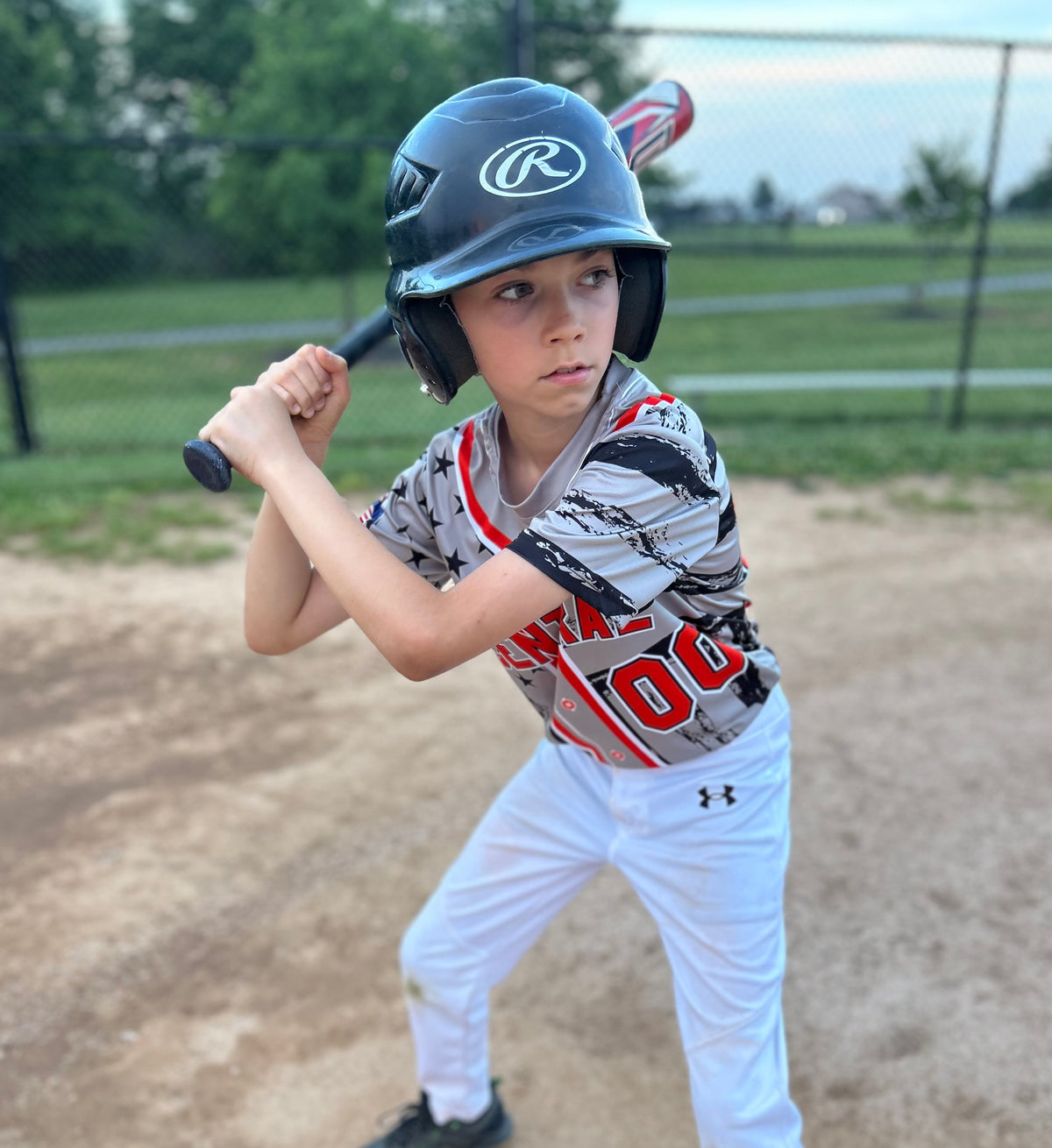 Young baseball player in a blue helmet and gray jersey with stars, standing on a dirt field
