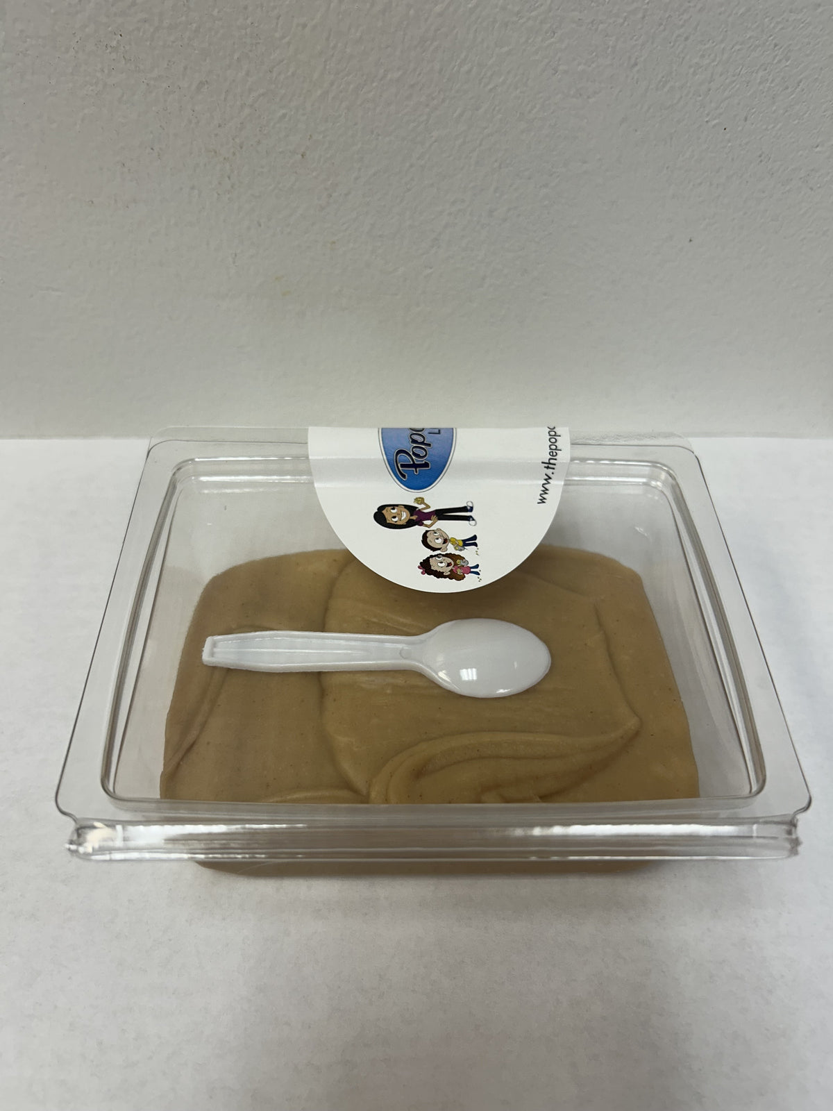A clear plastic clamshell package containing peanut butter fudge, with a small white plastic spoon resting on top and a white label sticker at the front.