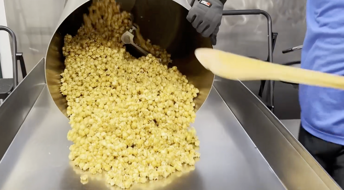 Popcorn being poured from a large metal hopper onto a conveyor belt, with a person in a blue shirt and gray glove visible on the right side.