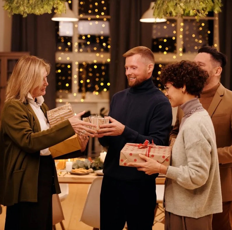 People at a warmly lit holiday gathering exchange wrapped gifts with red ribbon and dotted paper in the background