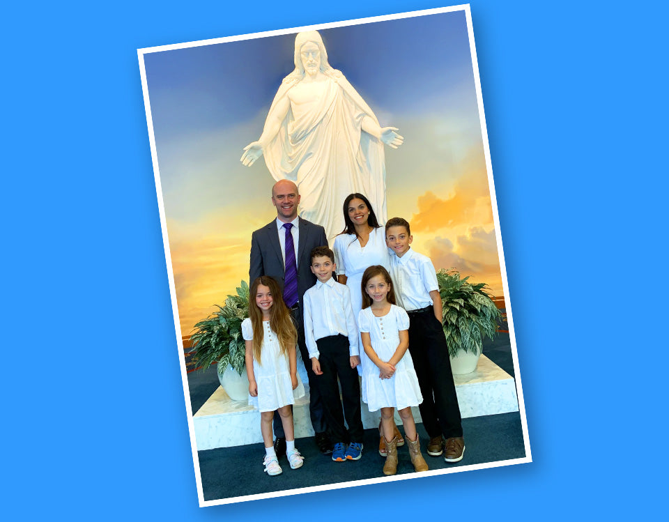 A family of seven poses for a photo in front of a large white statue of the Virgin Mary, with a sunset sky backdrop. The photo is shown as a tilted print or card against a solid blue background.