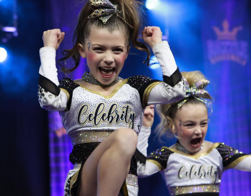 Two young girls in sparkling cheerleading outfits with 'Celebrity' on their shirts, performing on a stage.
